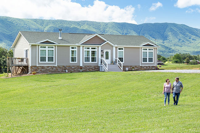 A couple walks across a spacious front yard of a beige manufactured home with lush mountains in the distance
