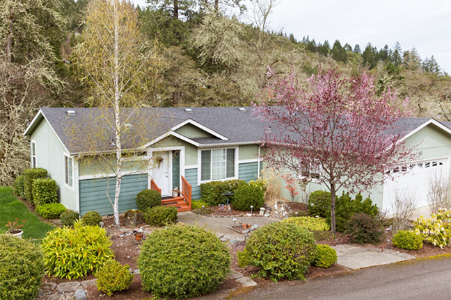 An elevated view of a manufactured home with a garage and planted shrubs, bushes, and trees