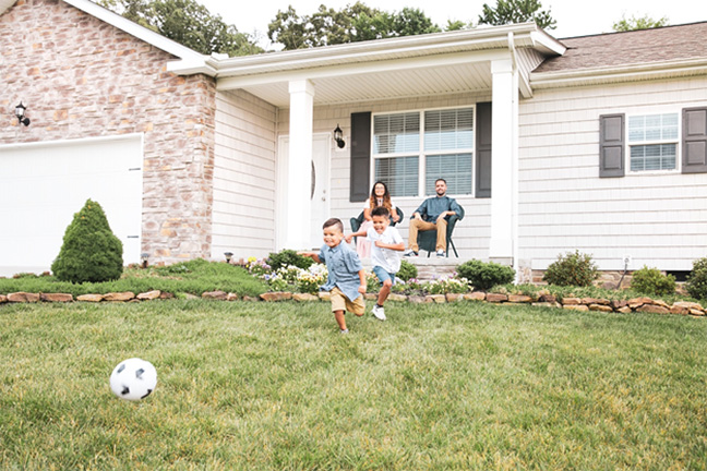 A husband and wife seated on the front porch watching their two boys chase a soccer ball in the yard.