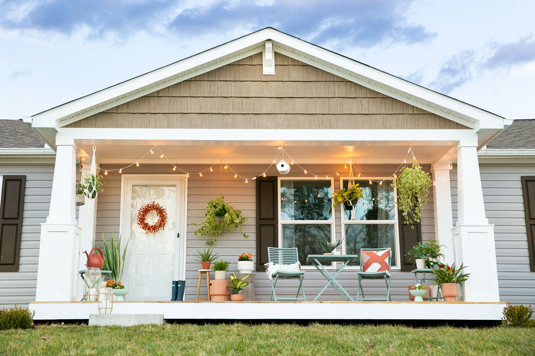A manufactured home pictured during the daytime. The home has several hanging plants, pieces of outdoor furniture, and decorative lights outside and looks very comfortable.