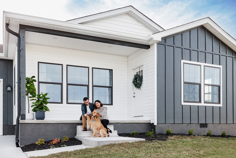A couple sits on the front porch of their manufactured home while petting their dog.