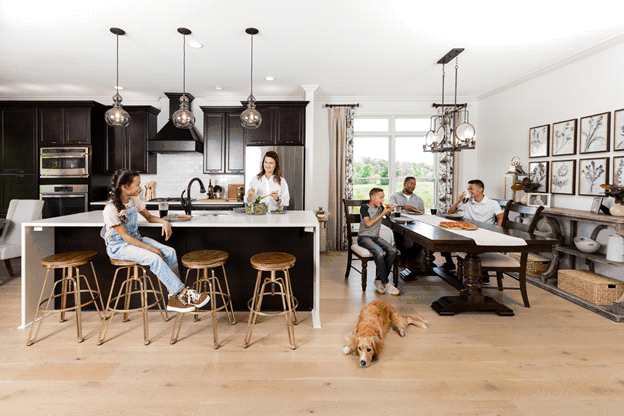 A family has gathered together in the kitchen of their new home. The kitchen is open-concept and spacious, with light pine board flooring and white walls. The father and two sons sit at a long dining table while mother and daughter are together at the kitchen counter. A dog is laying on the kitchen floor.