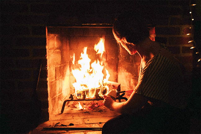 A woman in a striped blouse tends to a fire in a brick fireplace.
