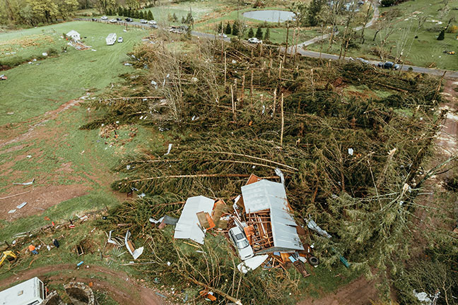 A heavily damaged manufactured home, surrounded by fallen trees and scattered debris.