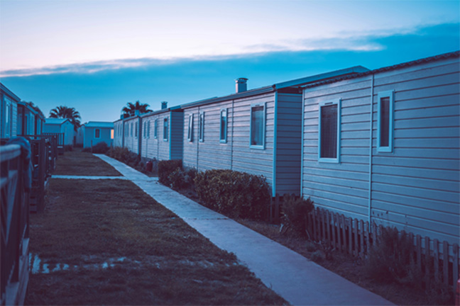 A cement walkway running between rows of mobile homes cast in shadow by the setting sun.