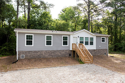 Gray manufactured home surrounded by towering trees.