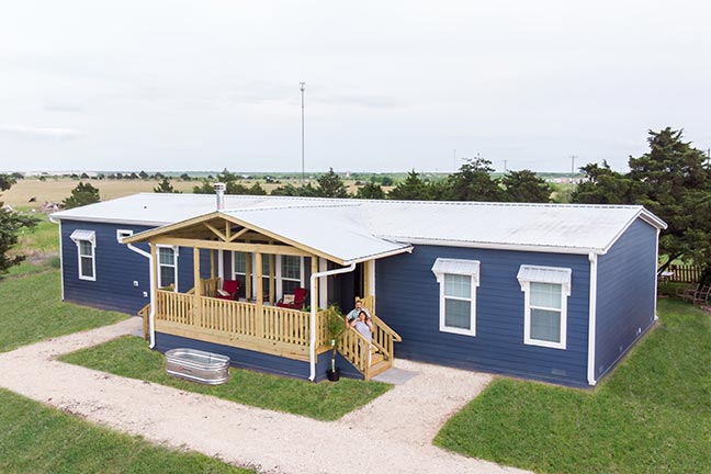 A couple stands on the steps of the wooden porch of their navy manufactured home, freshly mowed grass surrounding it.