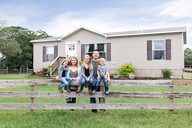 A man and woman stand behind their three daughters, who are sitting on the wooden fence surrounding their manufactured home.