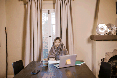 Woman bundled under a blanket in front of a window with thermal lined curtains to keep warm in her home