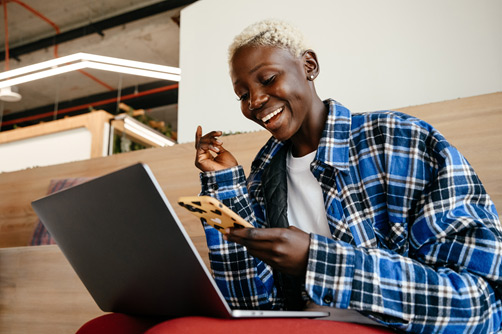 A short-haired black woman in a blue and black flannel stares happily at her computer screen upon realizing how much of a discount she is getting on her auto insurance policy.