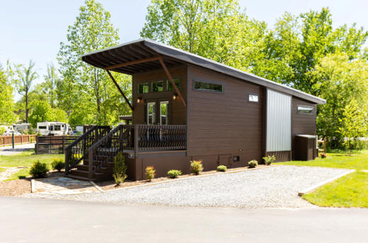 Simple brown stained wooden tiny home with metal roof sloping towards trees behind home.