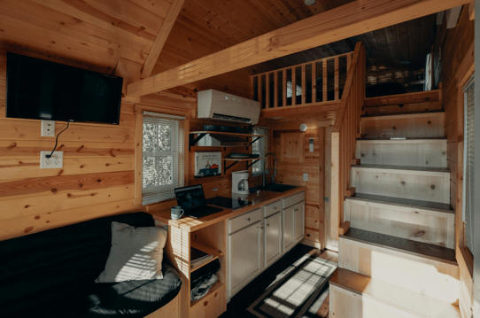 Cozy interior of a tiny home with exposed-wood walls, a staircase leading to a sleeping area above the simple kitchen.