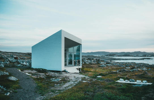 A modern tiny home sits on a rocky coastline, a large body of water a short distance away.