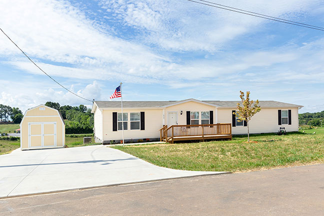 Cream colored mobile home with a matching shed at the end of a cement driveway, an American flag flying in the breeze in front of it.