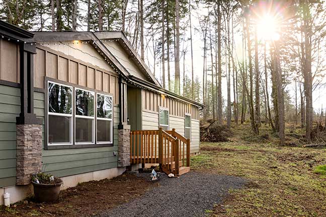 A mobile home with mint green siding and a small wooden porch sits in a clearing, surrounded by woods at sunset.