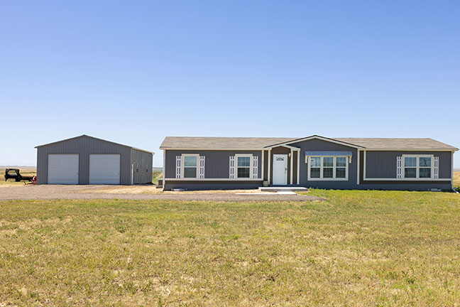 A gray mobile home with a matching detached garage, surrounded by dry grass and clear blue skies.